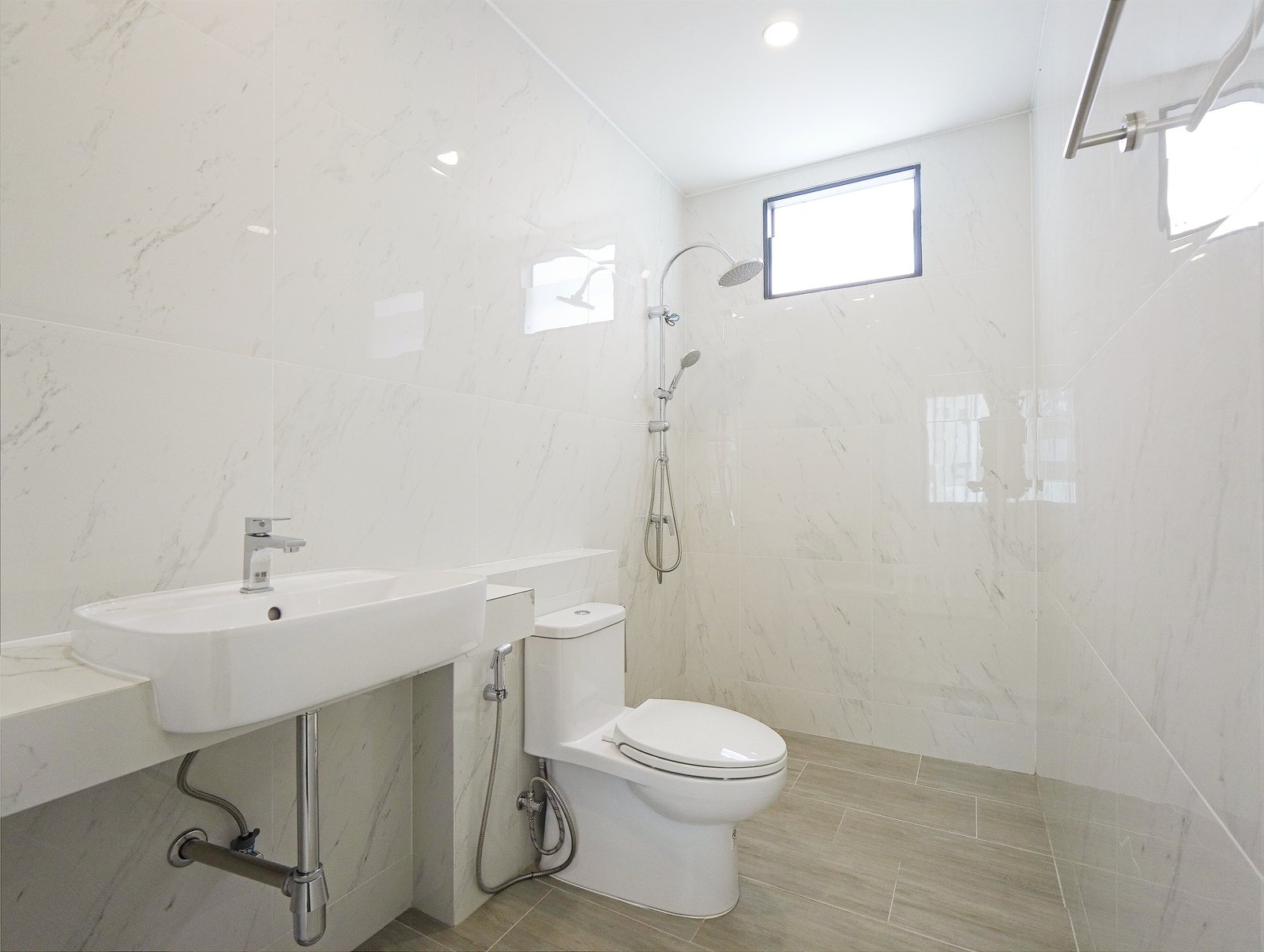Tropical_Village_3_Pattaya_Dlh453_8 White marble-tiled bathroom with a wall-mounted sink, toilet, and a modern shower area featuring a rainfall head and handheld sprayer, window on the far wall.