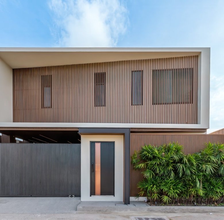 Modern two story house with wooden slat screen large entry door and tall black gate lush green bushes along the front right side