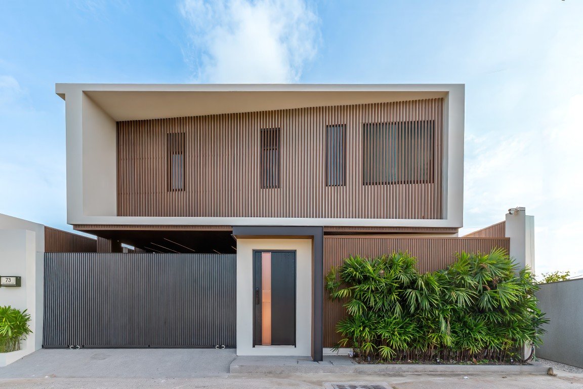 Modern two-story house with wooden slat screen, large entry door, and tall black gate; lush green bushes along the front right side.