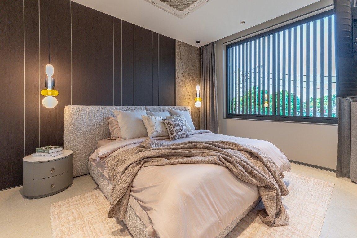 Modern bedroom with an upholstered bed, layered beige bedding, and pendant lights on each side by a dark wood paneled wall near a large window with vertical blinds.