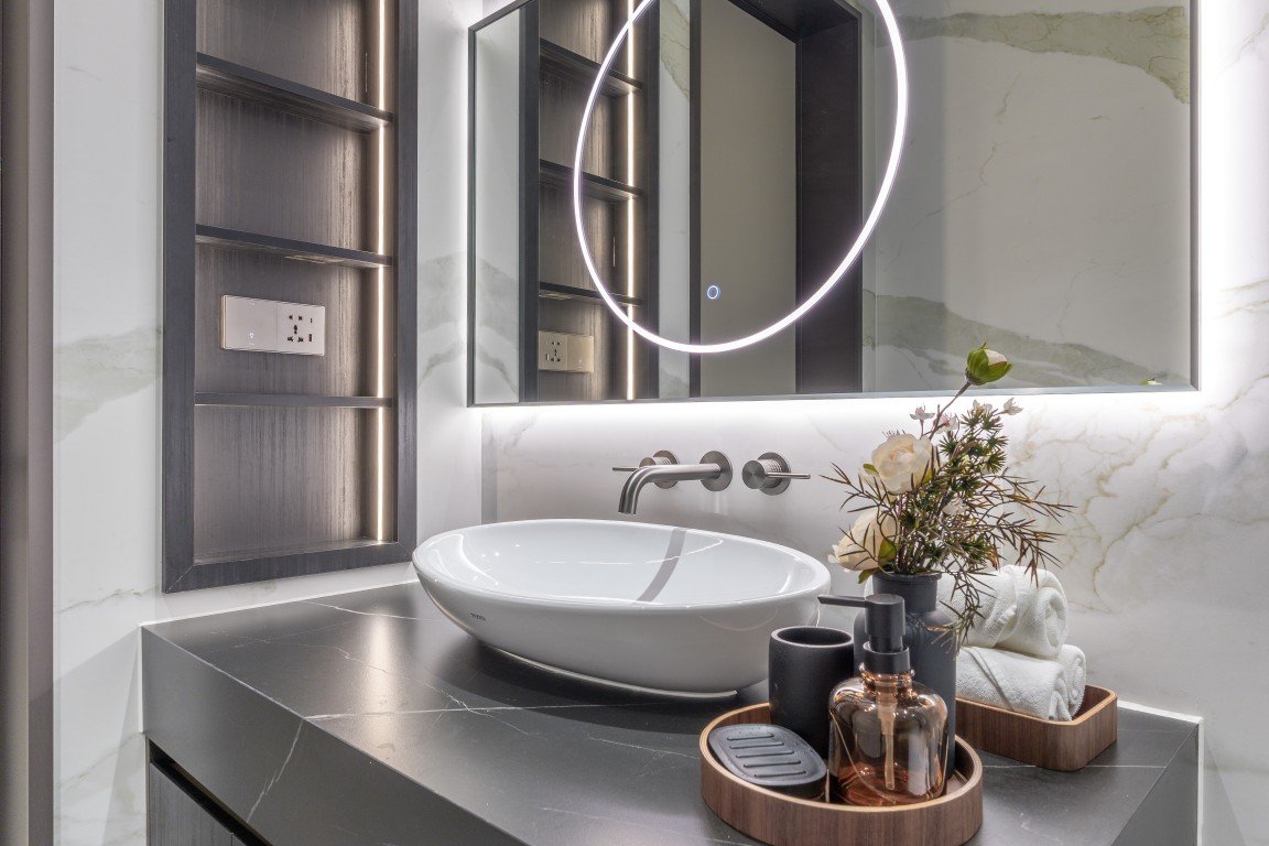 Modern bathroom vanity with a white vessel sink, wall-mounted faucet, backlit circular mirror, and neatly arranged toiletries on a tray.