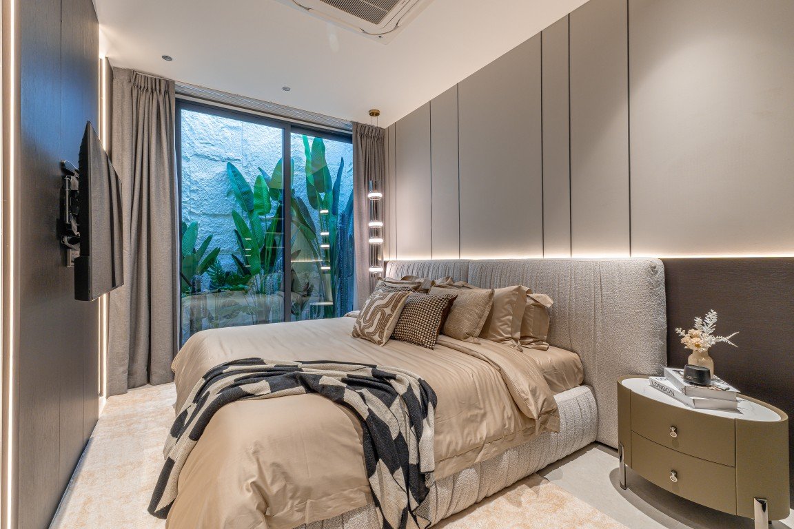 Luxurious bedroom with a large beige bed, tufted headboard, decorative pillows, and a striped throw; side table with books and vase on the right; floor-to-ceiling panels and soft ambient lighting.
