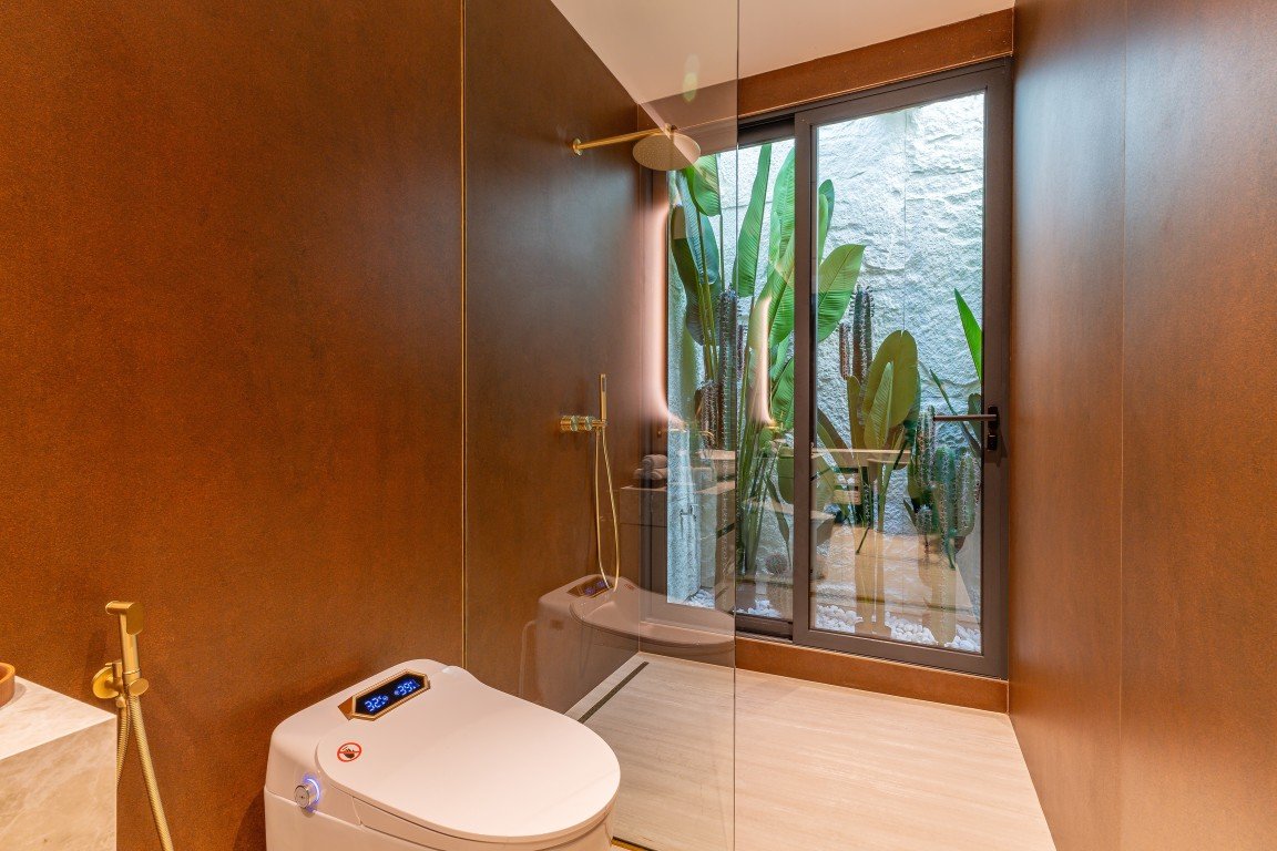 Modern bathroom with a white bidet-toilet, brown walls, and a glass door leading to a lush outdoor patio with tropical plants.
