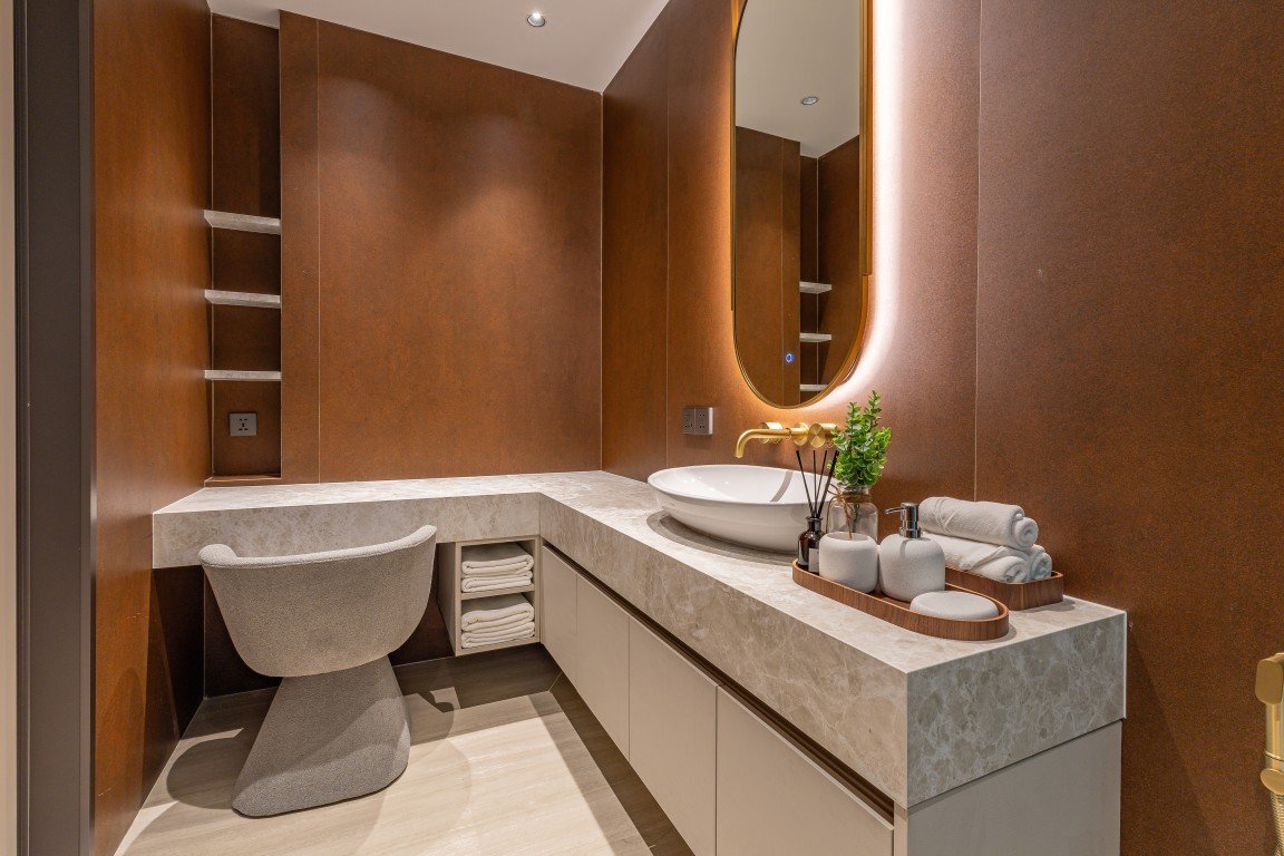 Contemporary bathroom with marble countertop, oval vessel sink, and backlit mirror; brown walls and a gray curved chair beside the vanity.