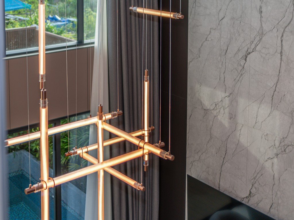 Contemporary pendant light with glowing orange tube segments hanging in front of a marble wall and dark curtains.