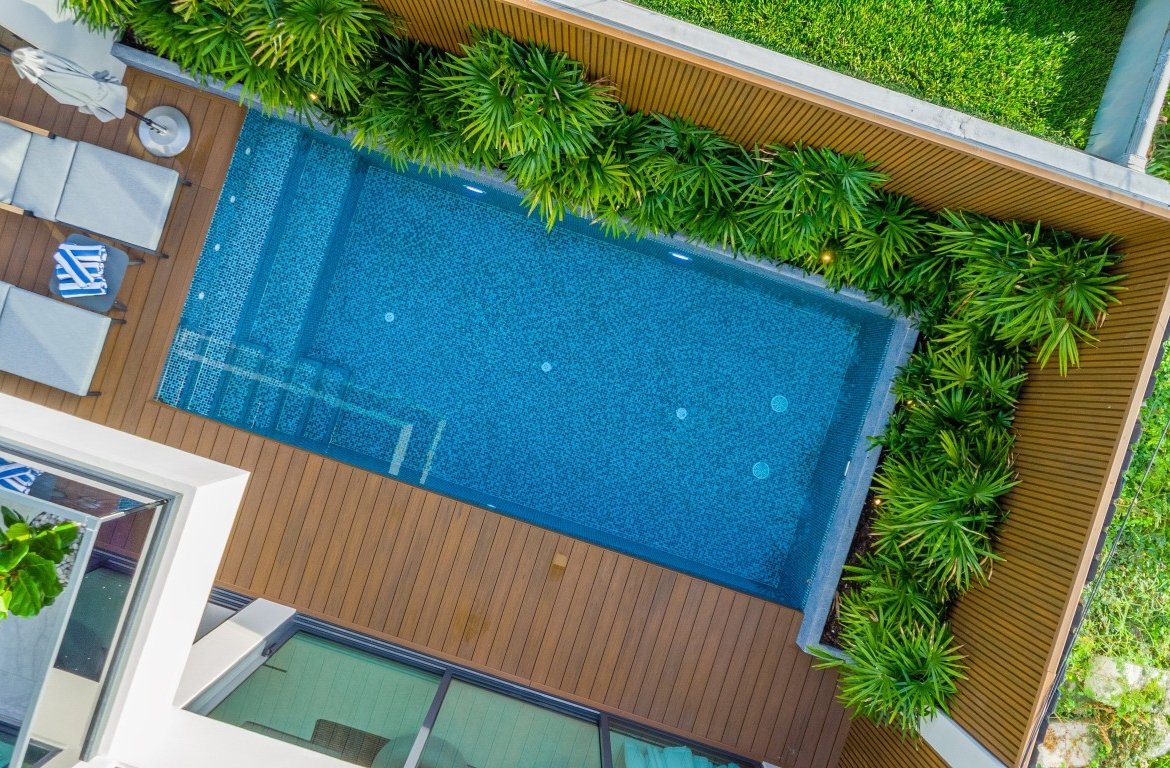 Aerial view of a rectangular blue-tiled pool surrounded by a wooden deck and lush green plants with lounge chairs nearby.