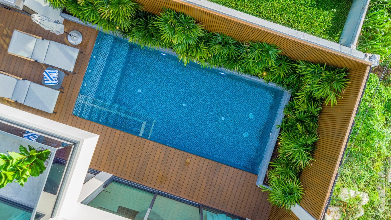 Villa_Solara_Pattaya_Picture1_45 Aerial view of a rectangular blue-tiled pool surrounded by a wooden deck and lush green plants with lounge chairs nearby.