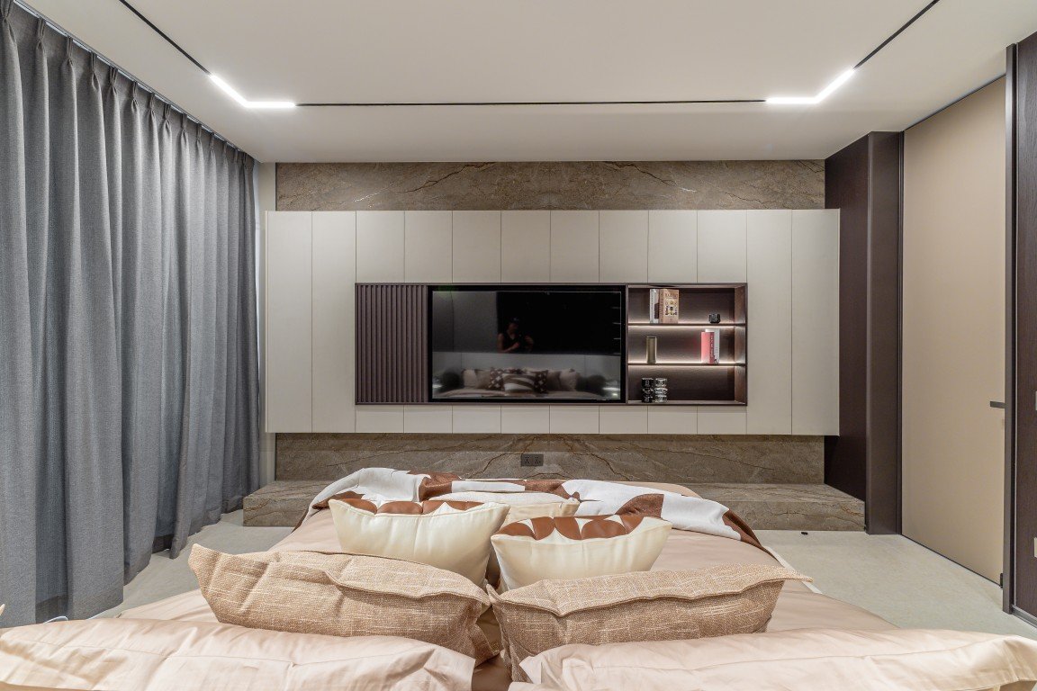 Modern bedroom with a wall-mounted TV unit, neutral cabinetry, and a plush bed in the foreground.