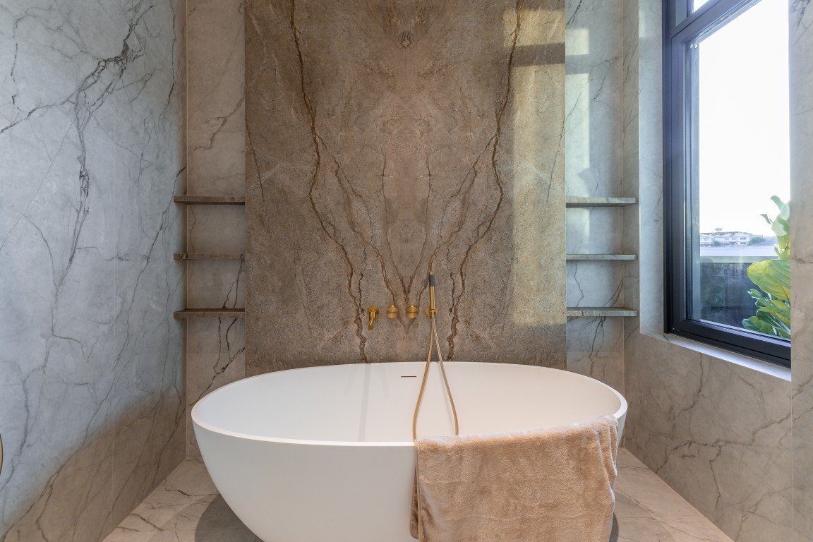 White freestanding bathtub with gold wall-mounted taps in a marble bathroom, with open shelves beside it and a window.