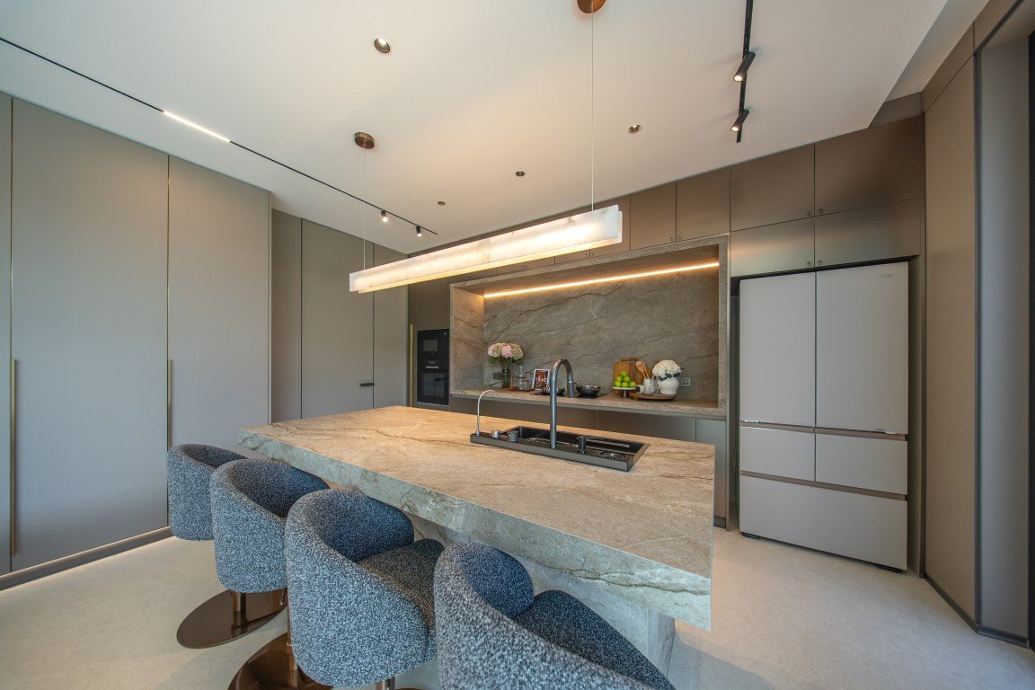 Contemporary kitchen with a long marble island, built-in sink, and gray cabinetry surrounding a stainless fridge.