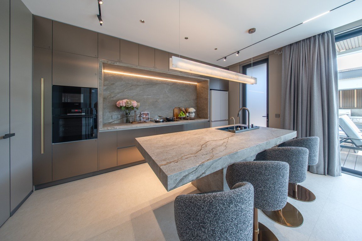 Modern kitchen with a large marble island and four grey upholstered bar stools, grey cabinets, built-in oven, and a window with curtains.