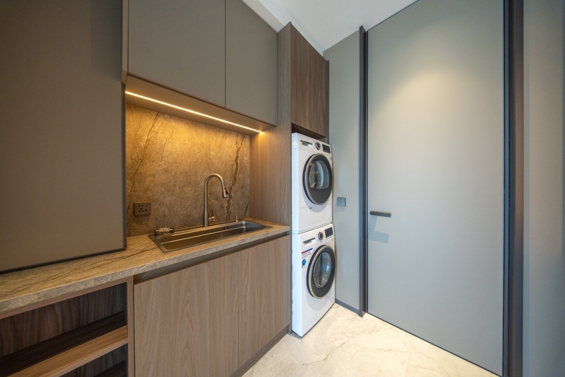 Modern laundry room with stacked front-load washer and dryer beside a gray door, wooden cabinets, and marble countertop.Microbreak: Opened to reflect the space accurately