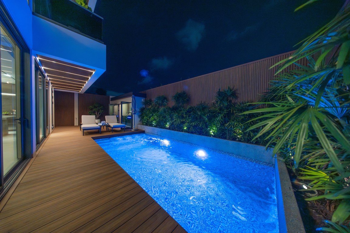 Nighttime pool area with blue-lit rectangular pool, wooden deck, and two lounge chairs on the deck near glass doors.