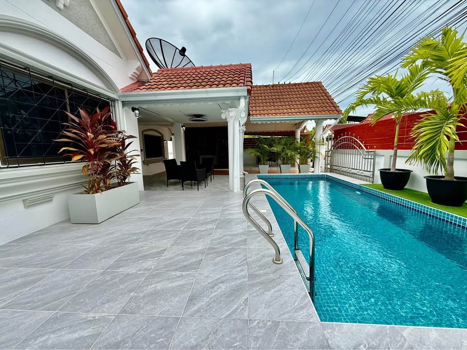 Backyard pool area with a gray tiled deck a covered patio and potted palm trees beside a blue rectangular pool