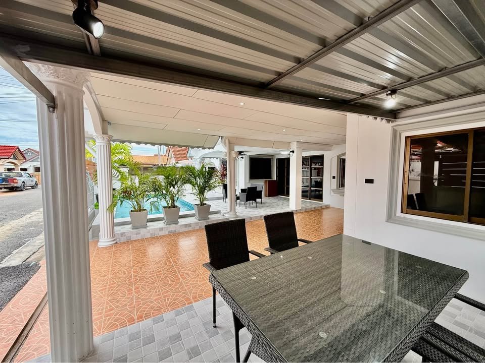 Covered outdoor patio with a glass dining table, four black wicker chairs, white columns, potted palms, and a pool beyond.