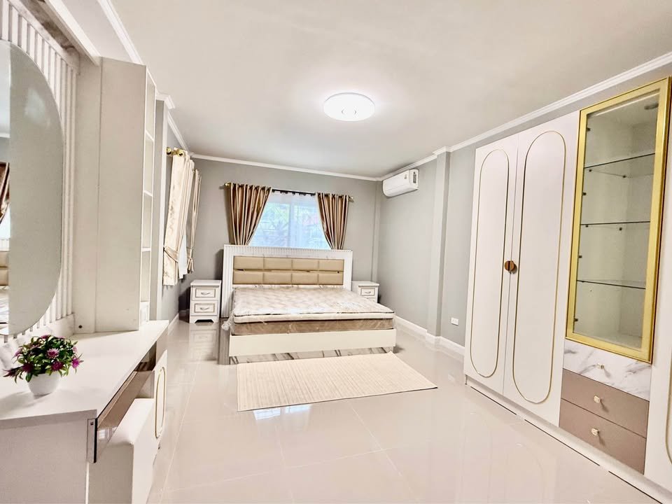 Bright modern bedroom with a beige upholstered bed, two white nightstands, a large white wardrobe, and cream curtains. A glass display cabinet and glossy tiled floor complete the scene.