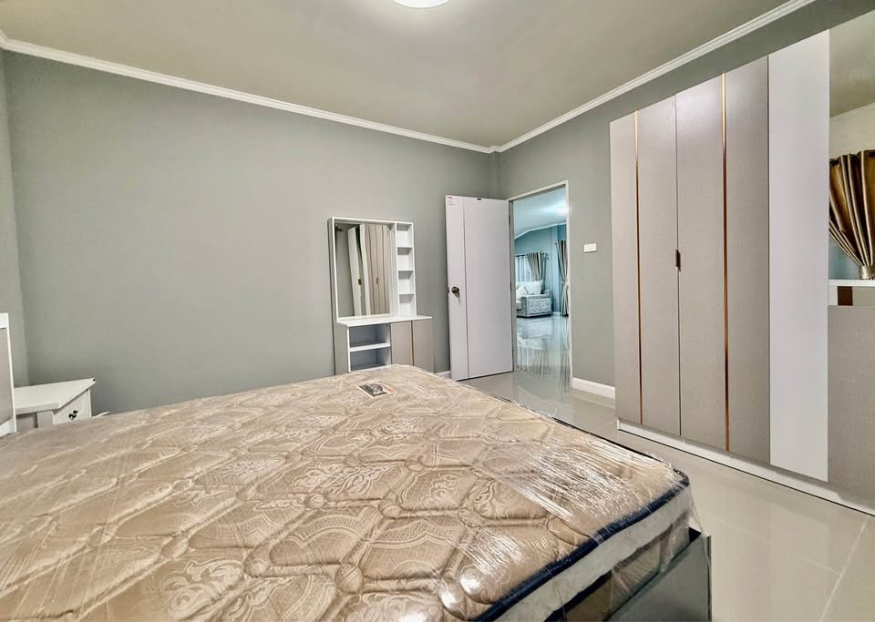 Bedroom with a large bed and patterned mattress, white side table, and a tall white wardrobe against gray walls; doorway to another room is open on the right.