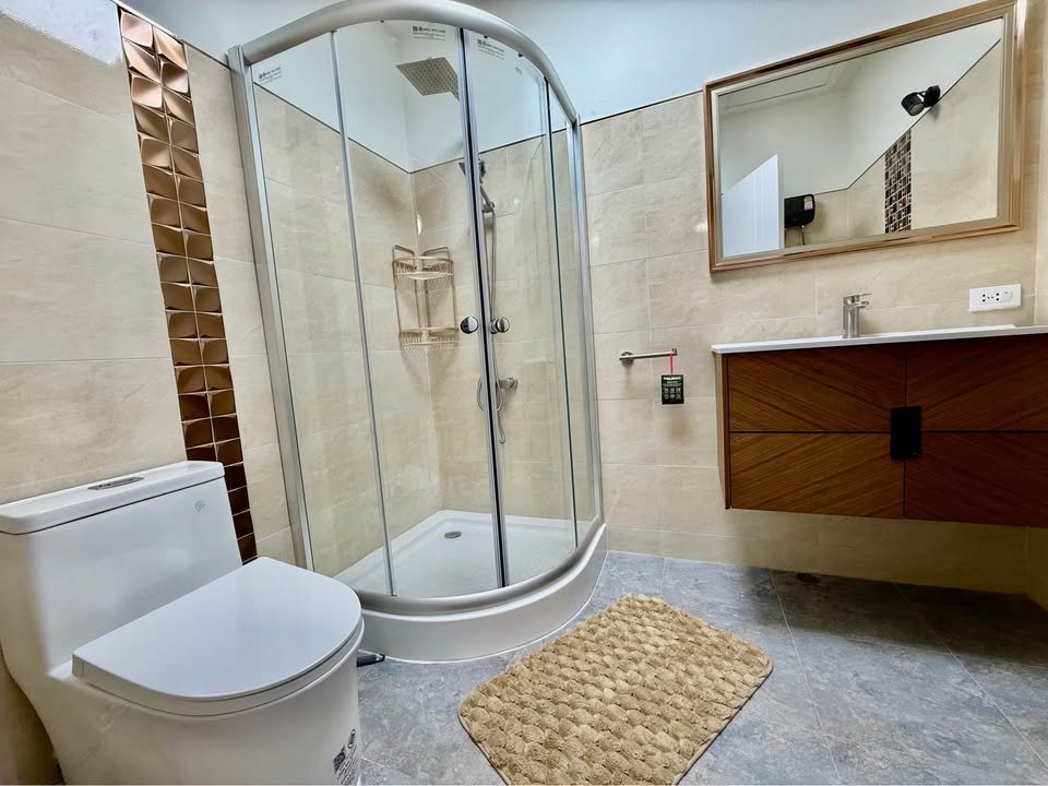 Modern bathroom with a curved glass shower enclosure, white toilet, and a wooden vanity under a mirror.