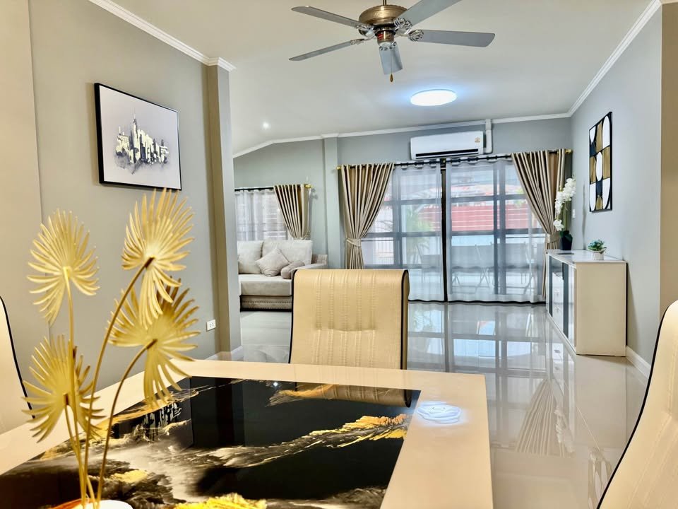 Open-concept dining and living room with a glass-top table and cream chairs, gold decorative plants, and large windows with sheer curtains; bright and modern.