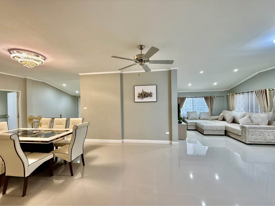 Open living and dining area featuring a ceiling fan, crystal chandelier, cream dining chairs, and a large beige sectional sofa in neutral tones.