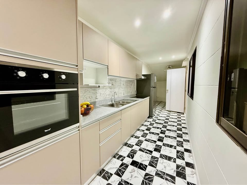 Modern galley kitchen with beige cabinets, built-in oven, stainless fridge, sink, and a black-and-white checkered floor.