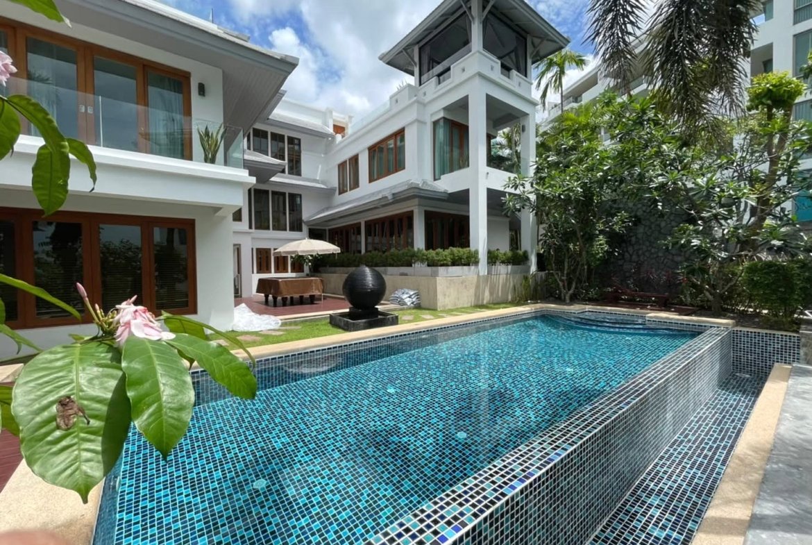 Luxurious white villa with glass balconies surrounding a rectangular blue-tiled pool, tropical plants nearby, under a partly cloudy sky.