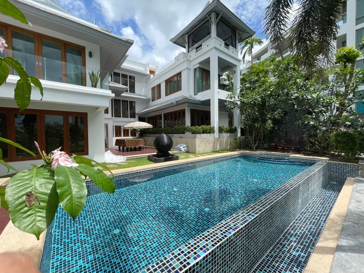 Luxurious white villa with glass balconies surrounding a rectangular blue-tiled pool, tropical plants nearby, under a partly cloudy sky.