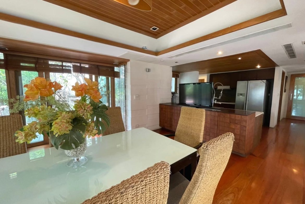 Dining room with a glass-top table, woven chairs, and a vibrant orange flower centerpiece; view toward the kitchen and a TV on a wooden cabinet.