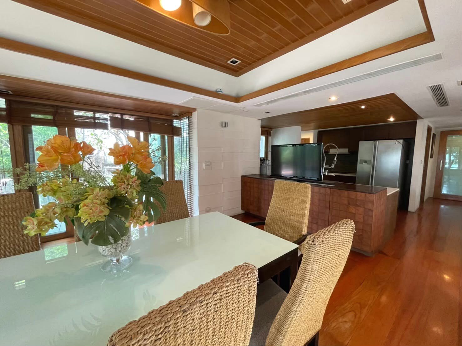 Dining room with a glass-top table, woven chairs, and a vibrant orange flower centerpiece; view toward the kitchen and a TV on a wooden cabinet.
