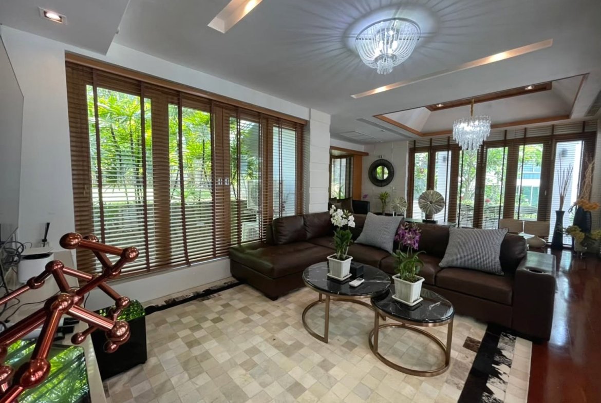Bright modern living room with a brown leather sectional, two round marble coffee tables, and large windows with wooden blinds.Outside greenery visible outside.