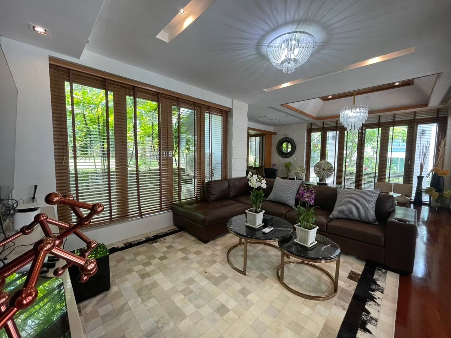Bright modern living room with a brown leather sectional, two round marble coffee tables, and large windows with wooden blinds.Outside greenery visible outside.