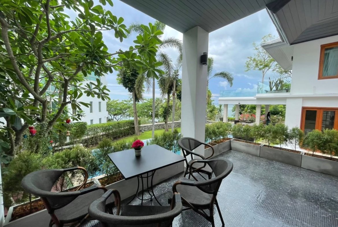 Outdoor balcony with a black metal table and four wicker chairs, pink flower centerpiece, looks out on a garden and pool area.