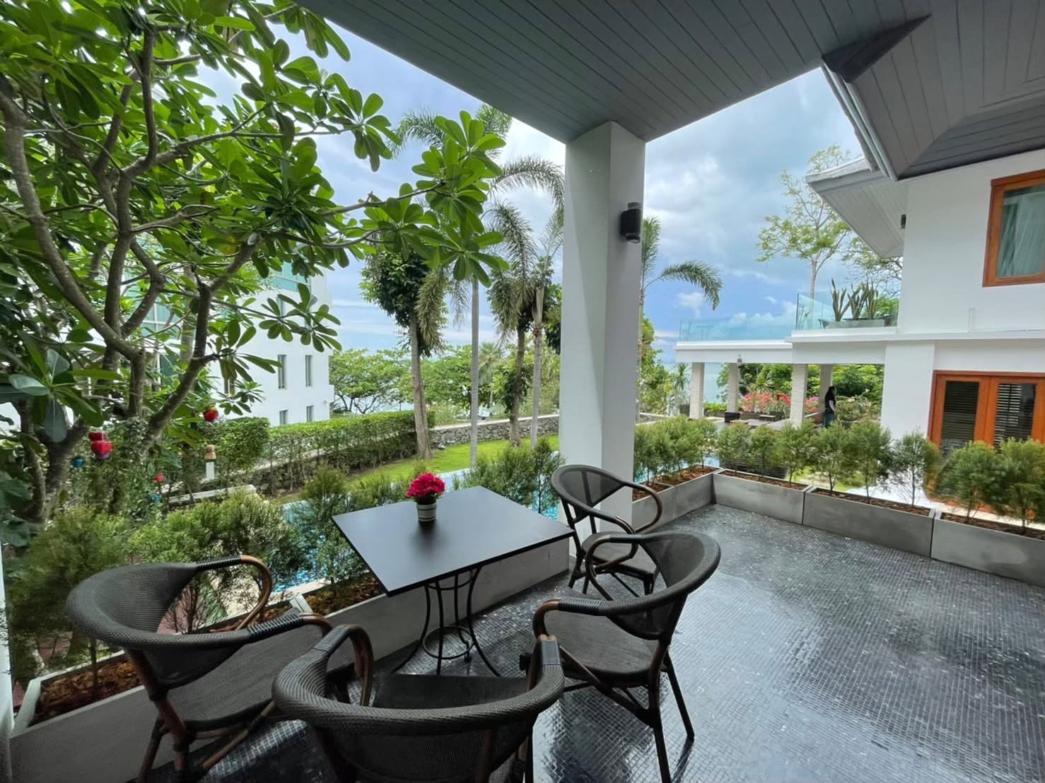 Outdoor balcony with a black metal table and four wicker chairs, pink flower centerpiece, looks out on a garden and pool area.