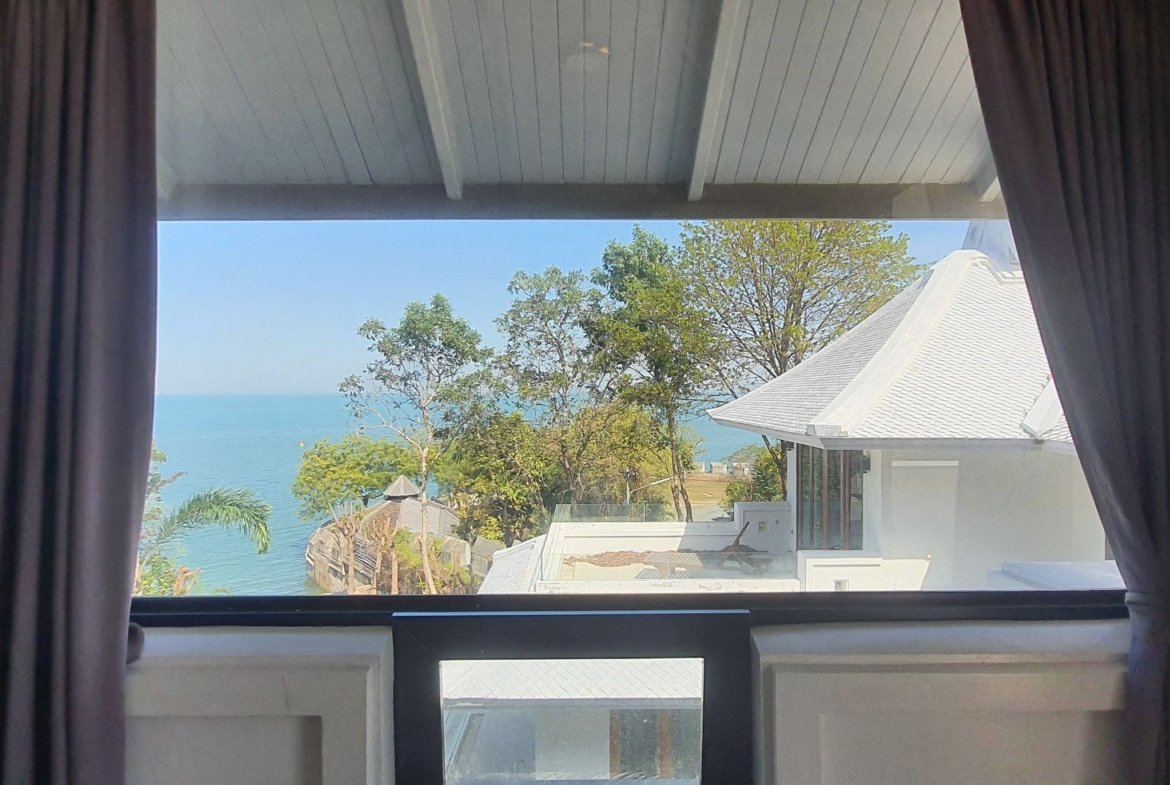 Window view of blue sea and sky framed by dark curtains, with white rooftops and greenery in the foreground.