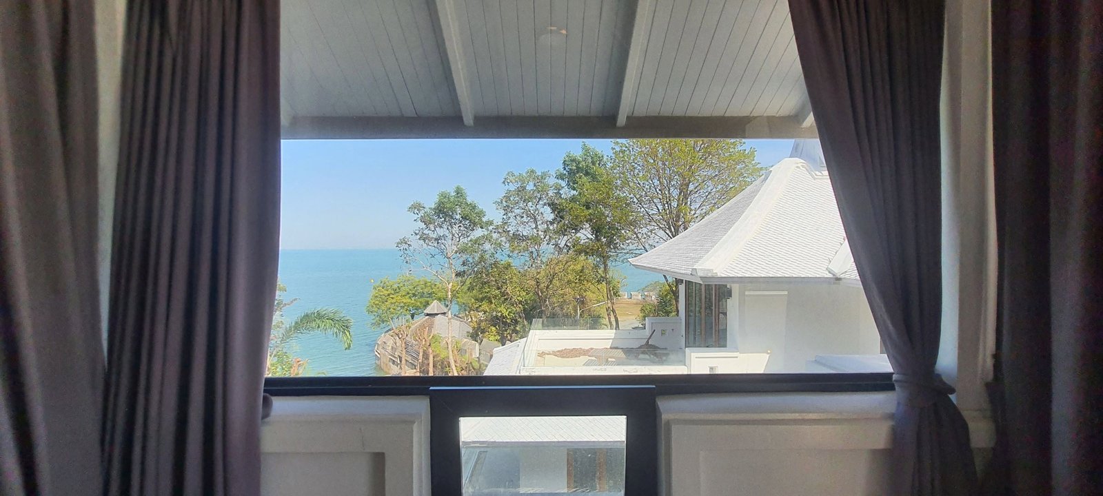 Window view of blue sea and sky framed by dark curtains, with white rooftops and greenery in the foreground.