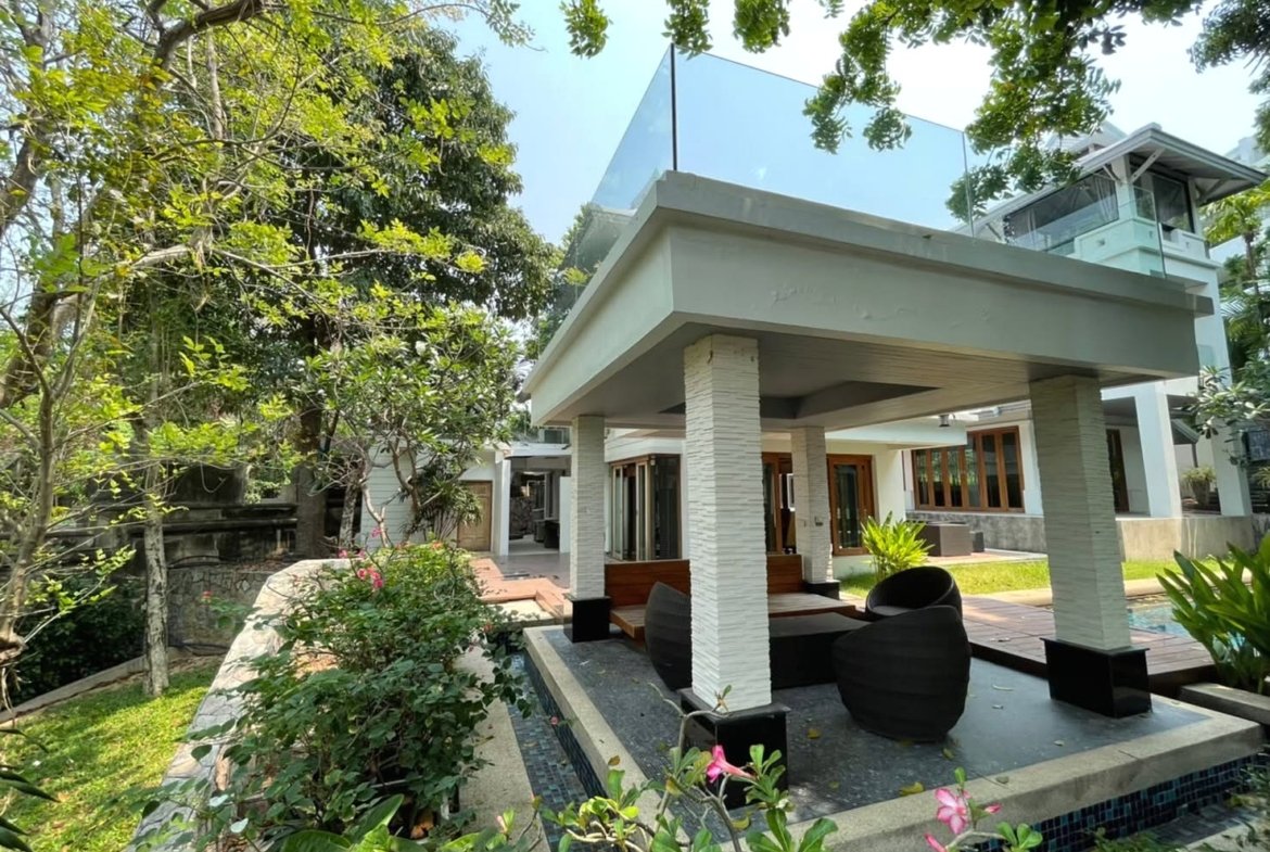Modern white villa with a covered patio, glass-railed second level, and tropical garden surroundings.