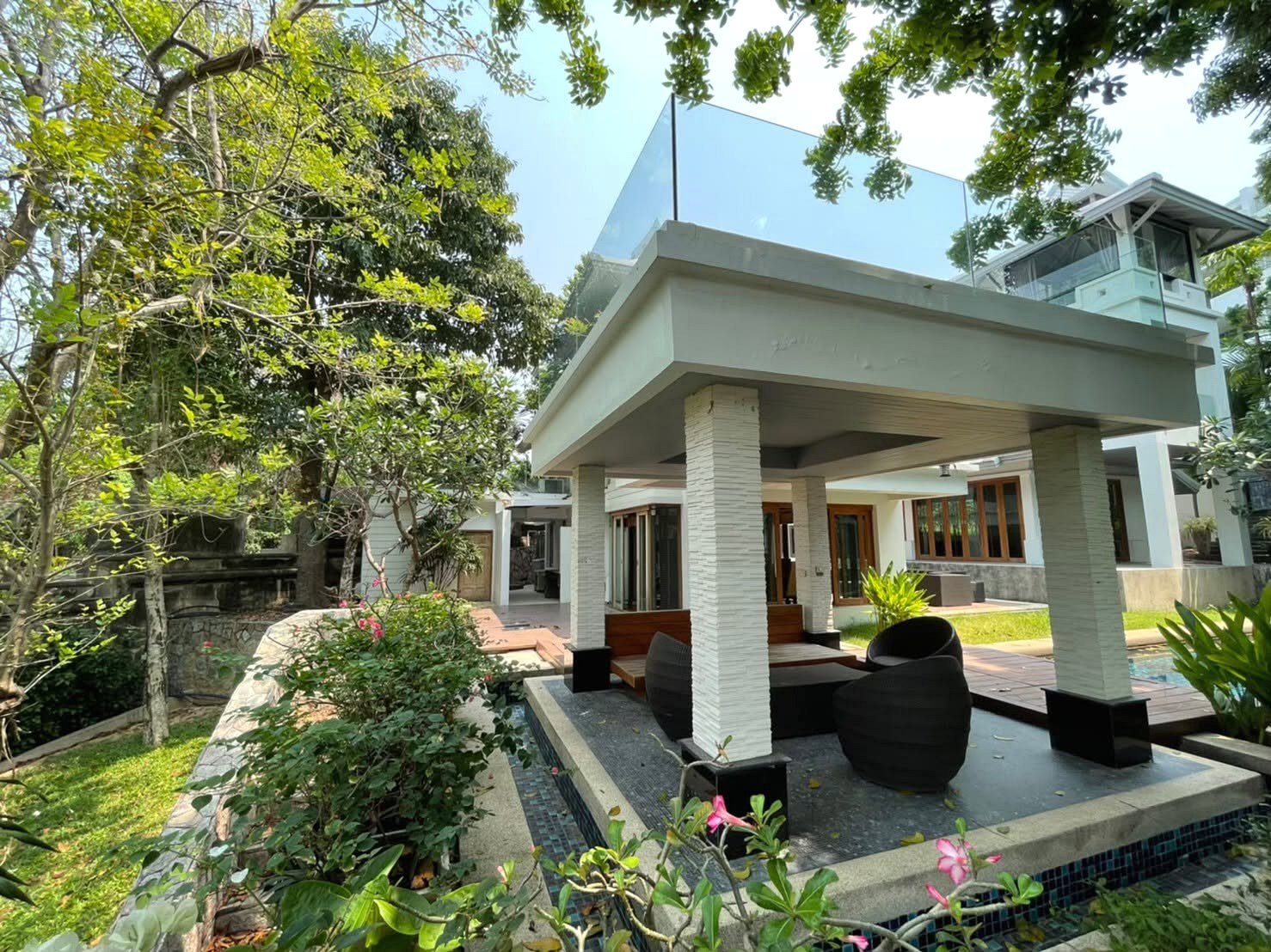 Modern white villa with a covered patio, glass-railed second level, and tropical garden surroundings.
