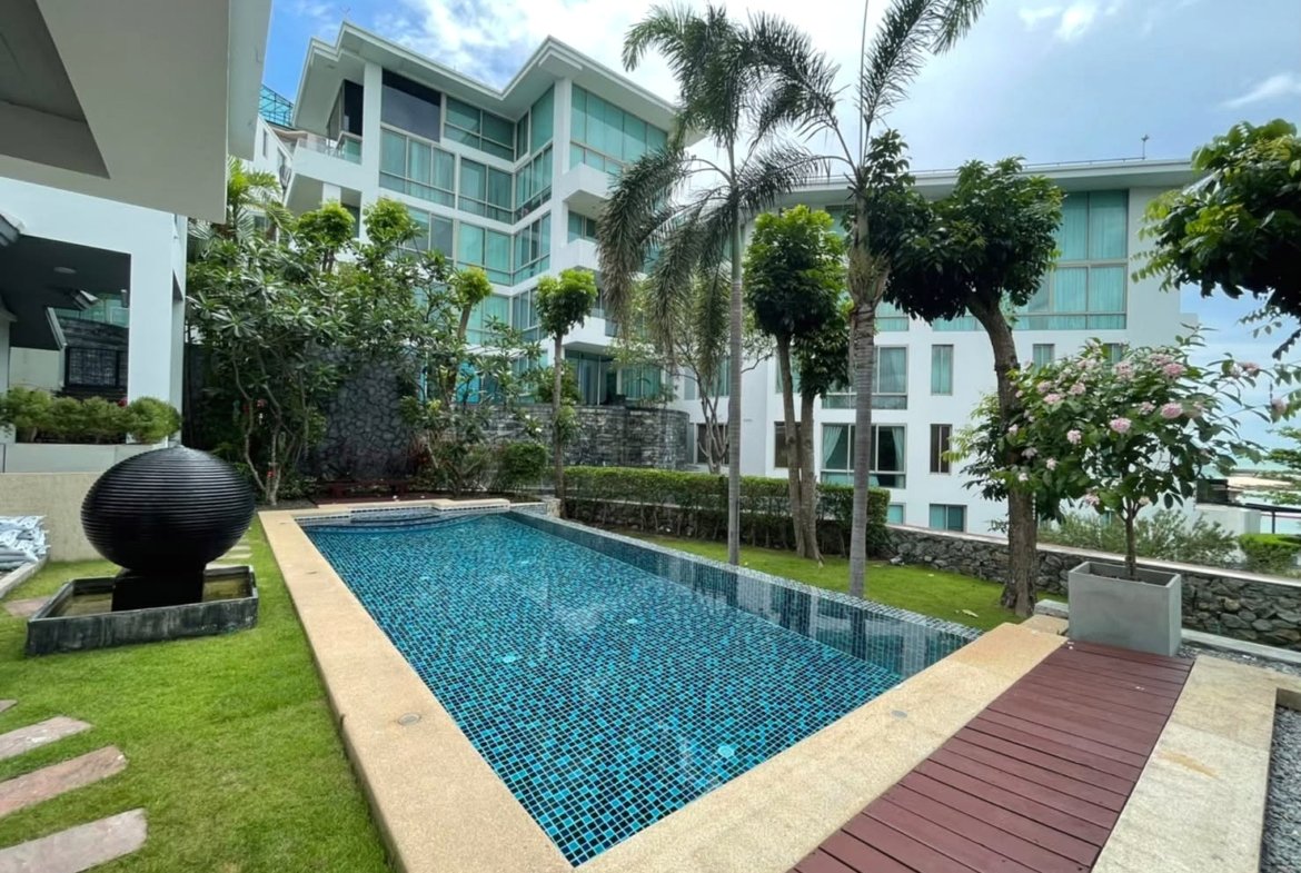 Resort-style courtyard with a rectangular blue-tiled pool, palm trees, and a white modern apartment building in the background.