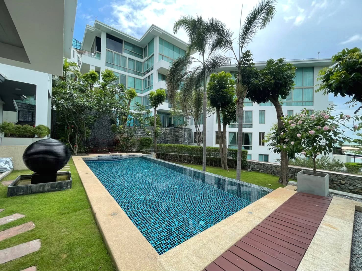 Resort-style courtyard with a rectangular blue-tiled pool, palm trees, and a white modern apartment building in the background.