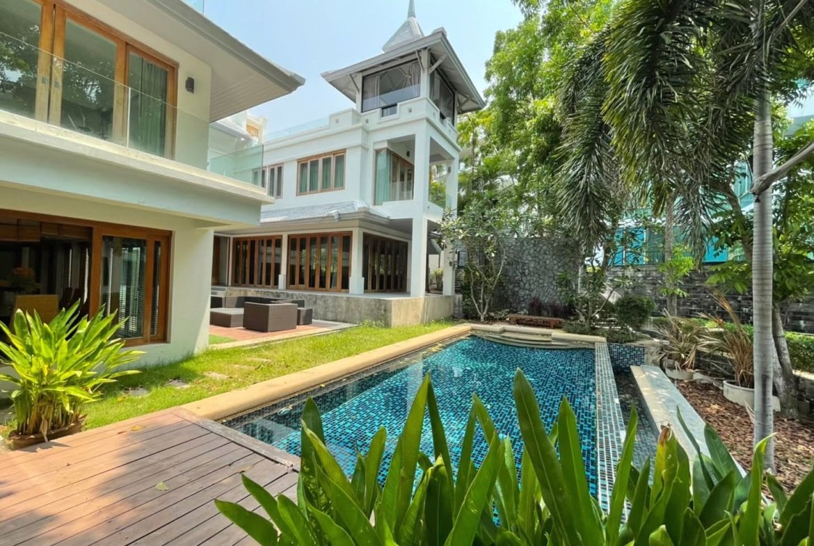 Modern white villa with glass balcony railings, a rectangular blue-tiled pool, wooden deck, and tropical plants.