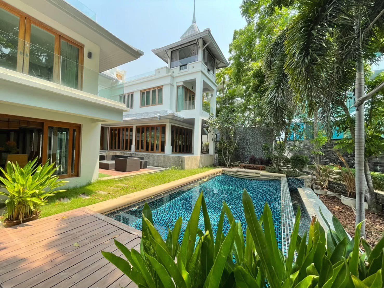 Modern white villa with glass balcony railings, a rectangular blue-tiled pool, wooden deck, and tropical plants.