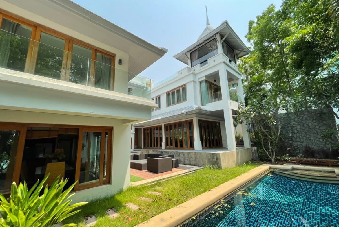 Modern white villa with glass railings, wooden-framed windows, and a blue-tiled pool in a sunny garden.