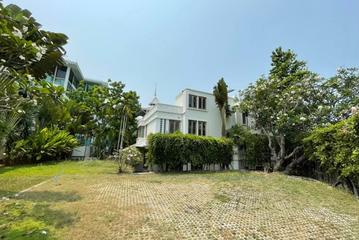 White two-story house with many windows, framed by lush tropical plants and a hedge under a clear blue sky.