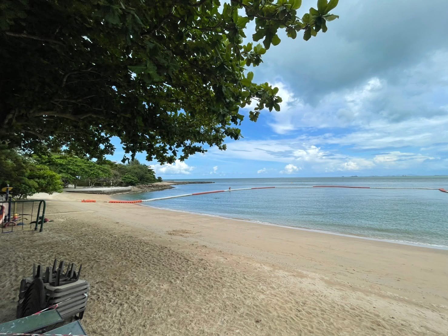 Beach scene with a large overhanging tree on the left, sandy shore, calm blue water, and a floating barrier extending into the sea under a partly cloudy sky.
