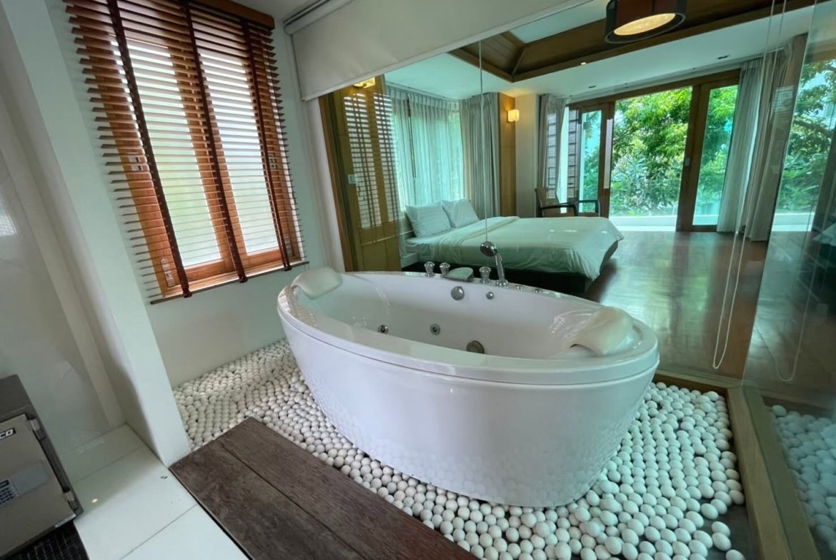 Luxurious bathroom with an oval Jacuzzi tub on a pebble floor, wood blinds, and a glass wall opening to a bedroom with a king bed and greenery outside.