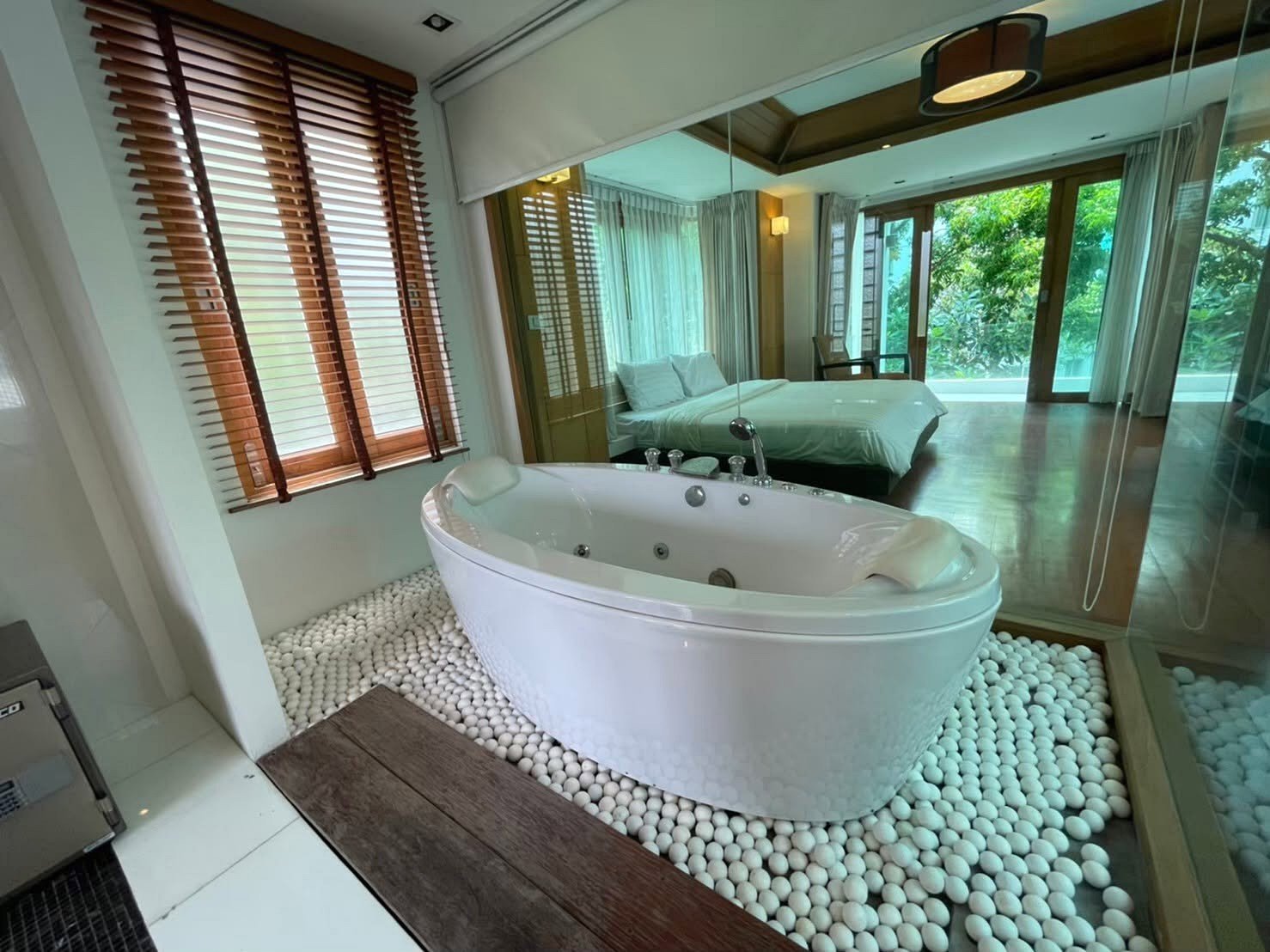Luxurious bathroom with an oval Jacuzzi tub on a pebble floor, wood blinds, and a glass wall opening to a bedroom with a king bed and greenery outside.