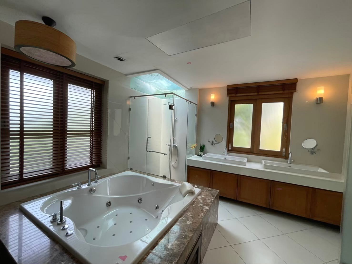 Luxurious bathroom with a large jetted tub in the foreground, a glass-enclosed shower to the left, and a double sink vanity under a wooden-framed window.