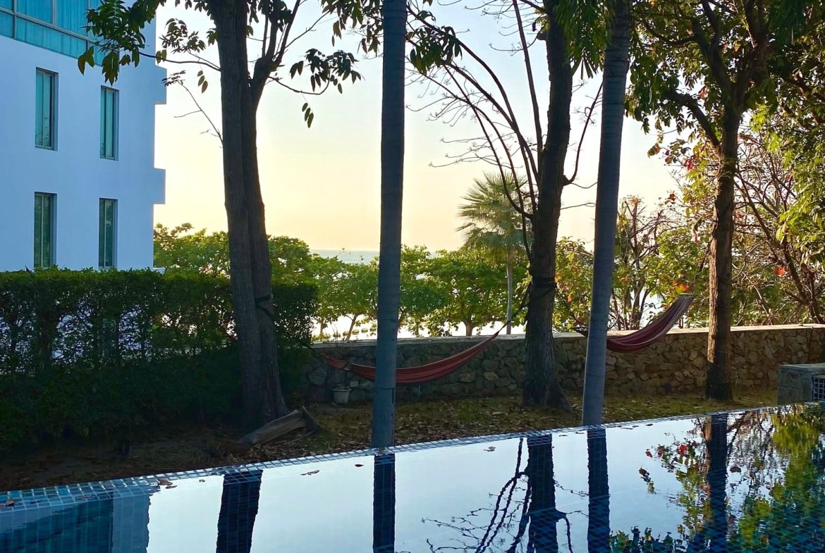Outdoor pool with blue-tiled surface reflecting surrounding trees and sky; a building with blue-tinted windows to the left and a hammock in the background.