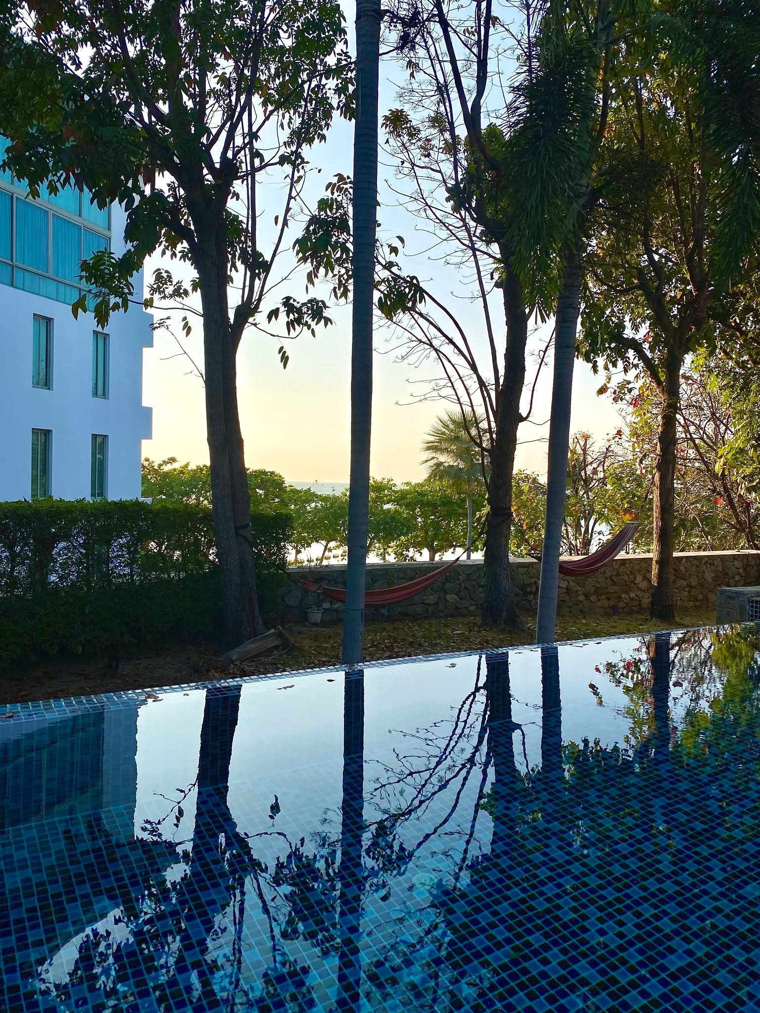 Wongamat_House_Rent_Rdl357_10 Outdoor pool with blue-tiled surface reflecting surrounding trees and sky; a building with blue-tinted windows to the left and a hammock in the background.