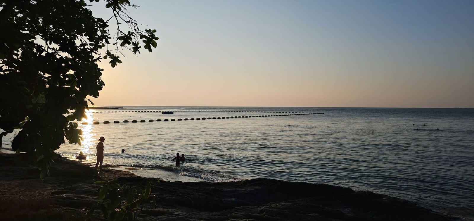 Wongamat_House_Rent_Rdl357_11 Sunset beach scene with a silhouetted tree on the left, calm water, and people swimming near shore as sun reflects off the sea.